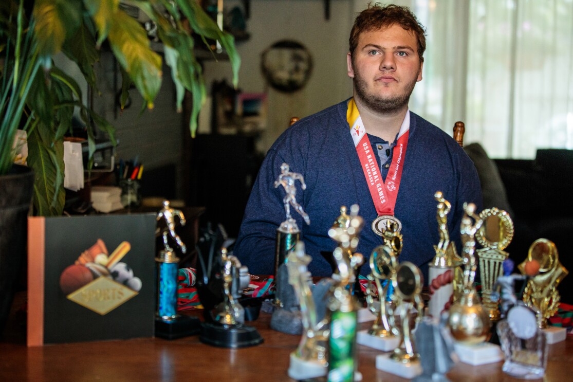 A man in a blue long-sleeved shirt wears a medal. In the foreground are many trophies.