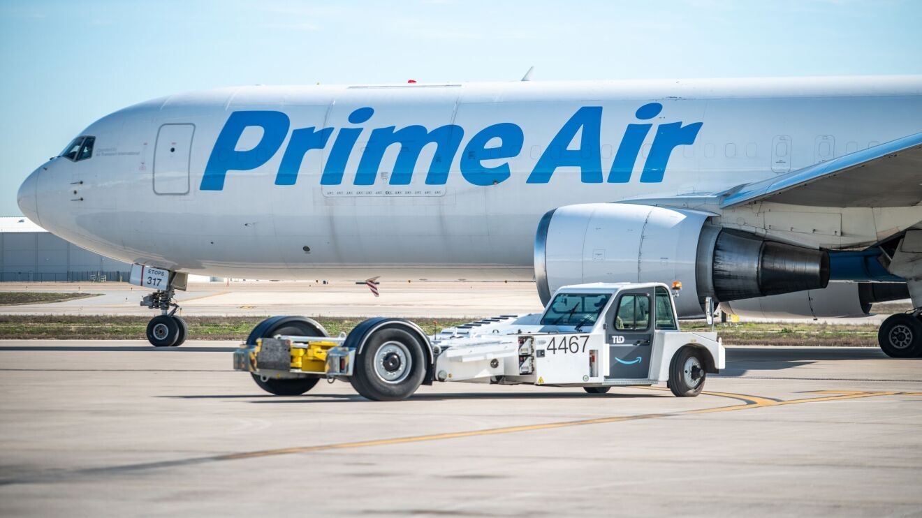 A photo showing half of an Amazon Air plane with a long, flat vehicle parked in front of it. The vehicle will eventually be used to push the plane back onto the taxiway.