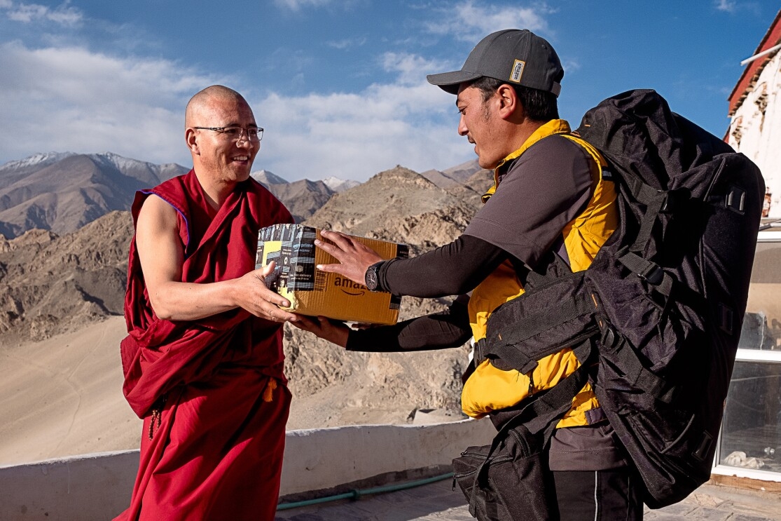 A delivery driver hands an Amazon package to a monk. Behind them, mountains and blue skies are visible.