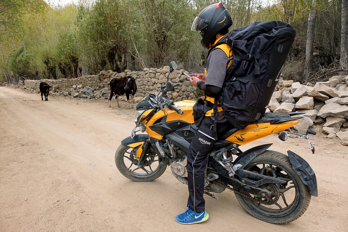 A motorcyclist wearing a large black backpack and an Amazon delivery uniform stands astride a motorcycle on a dirt road. He looks down and checks his smartphone. Two cows are in the background of the image.