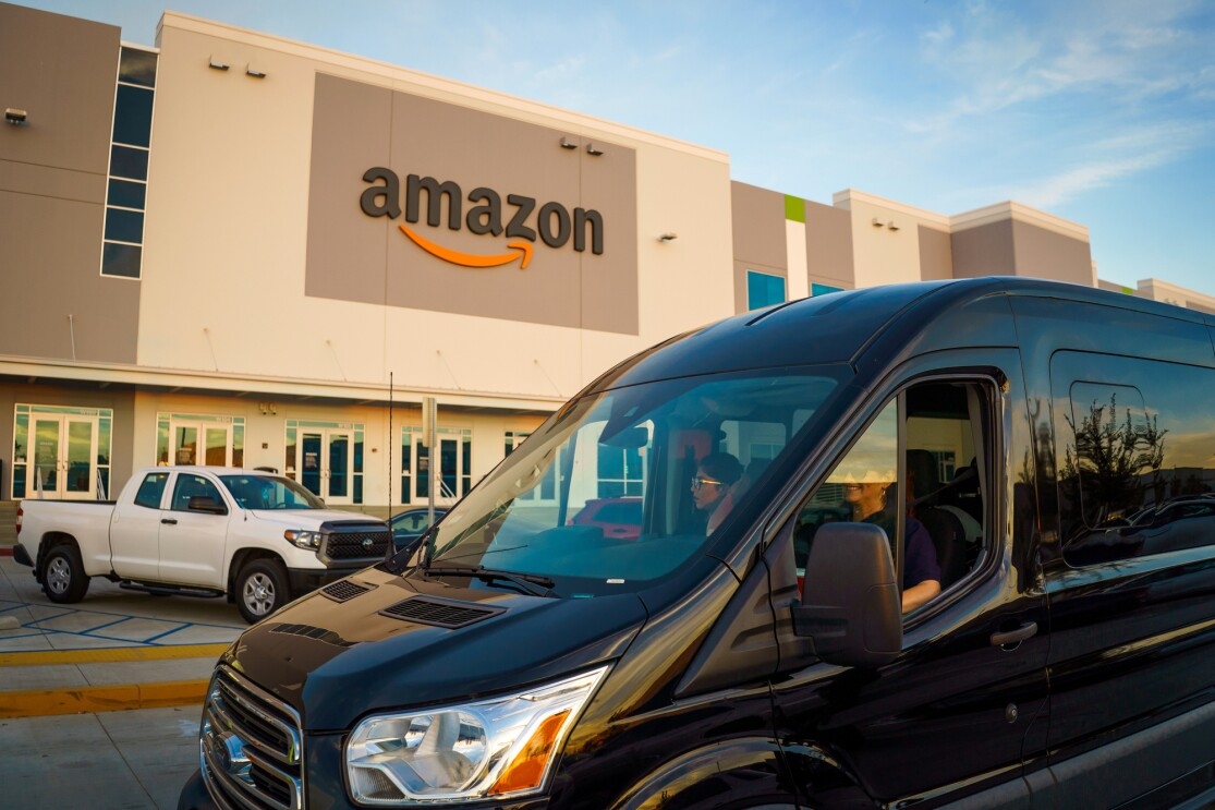 A shiny black van drives through a parking lot flanked by a building bearing the Amazon logo.