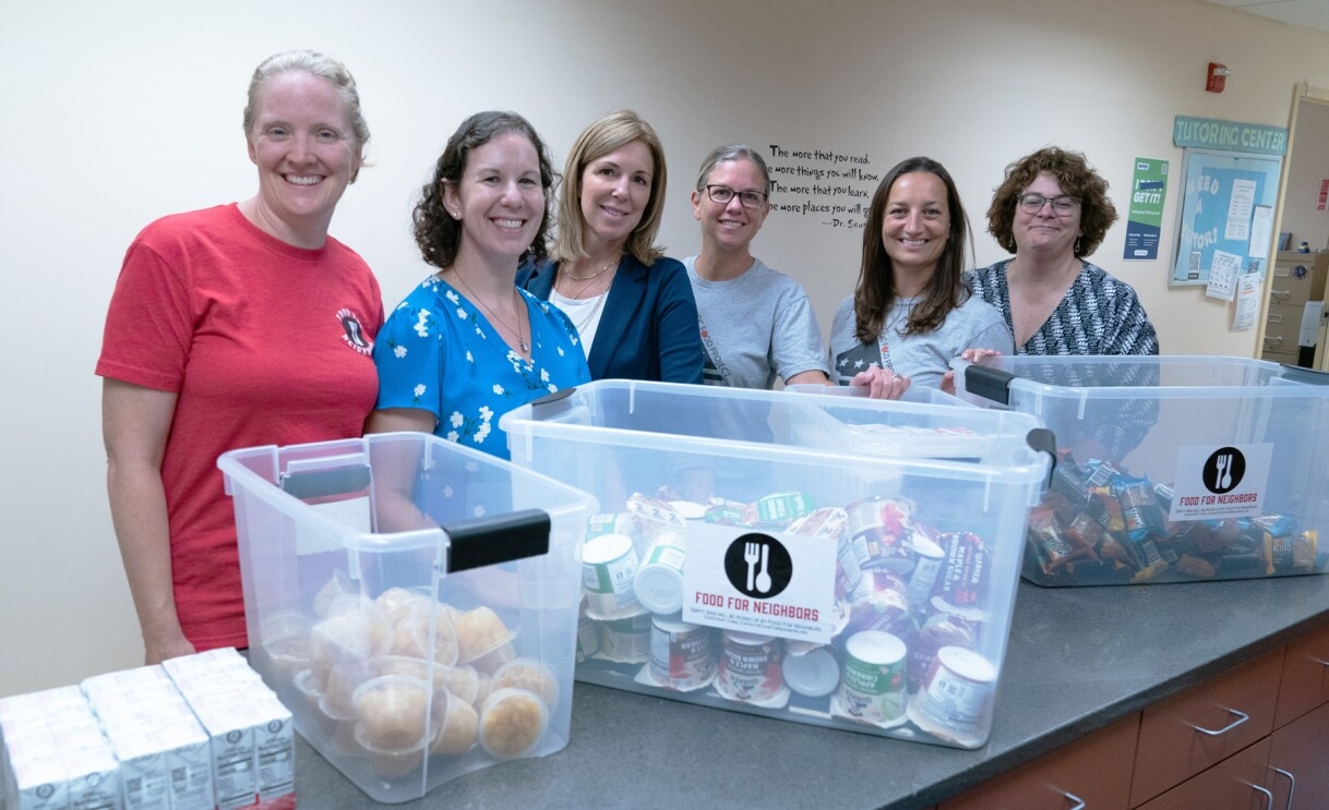 An image of six women volunteering to donate food to a school. There are three buckets of assorted food in front of them. 