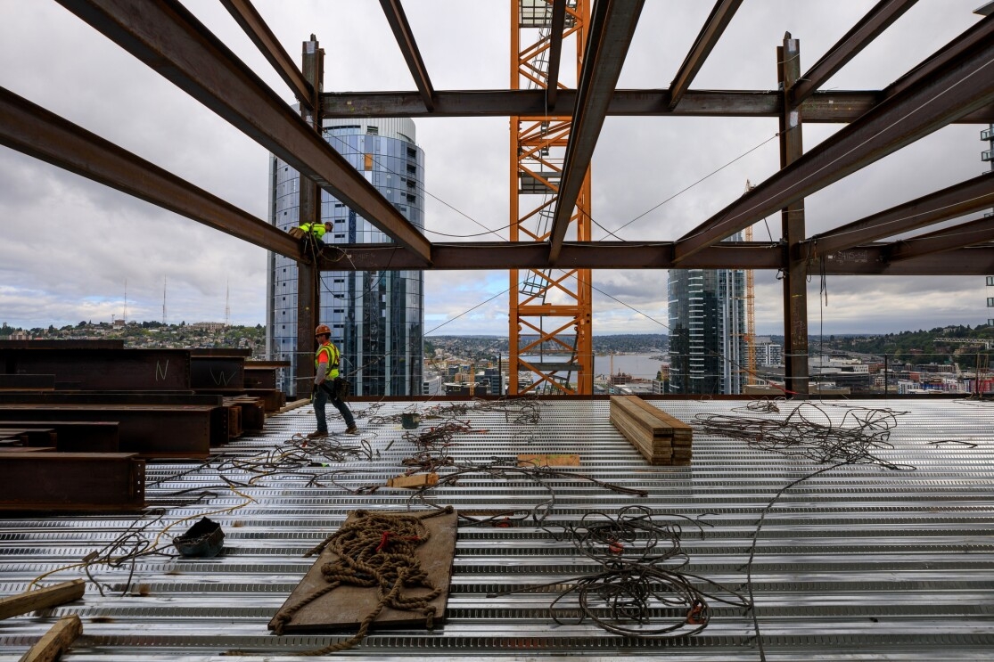 A construction worker walks across an under-construction Amazon skyscraper, with wires and support beams ready to be installed. Above him, another construction worker is on a steel support beam. 