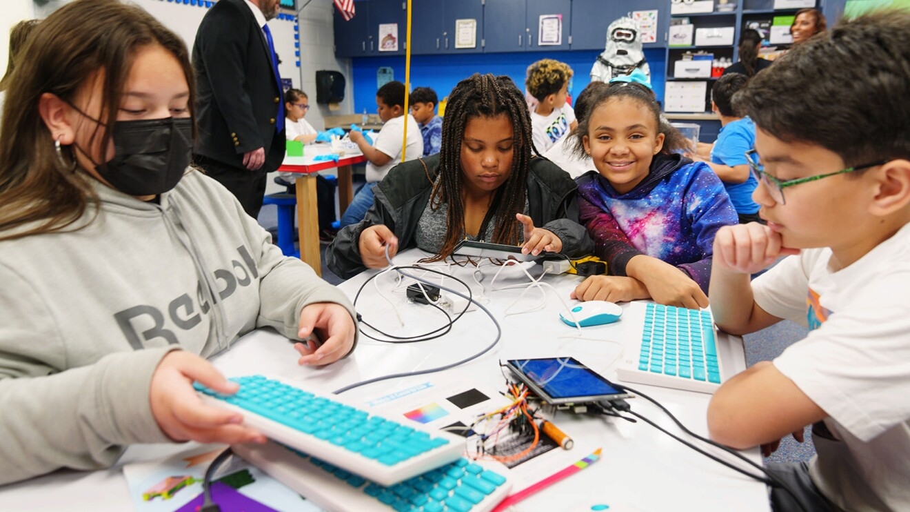 A photo of four students sitting at a table of electronics such as keyboards and tablets.