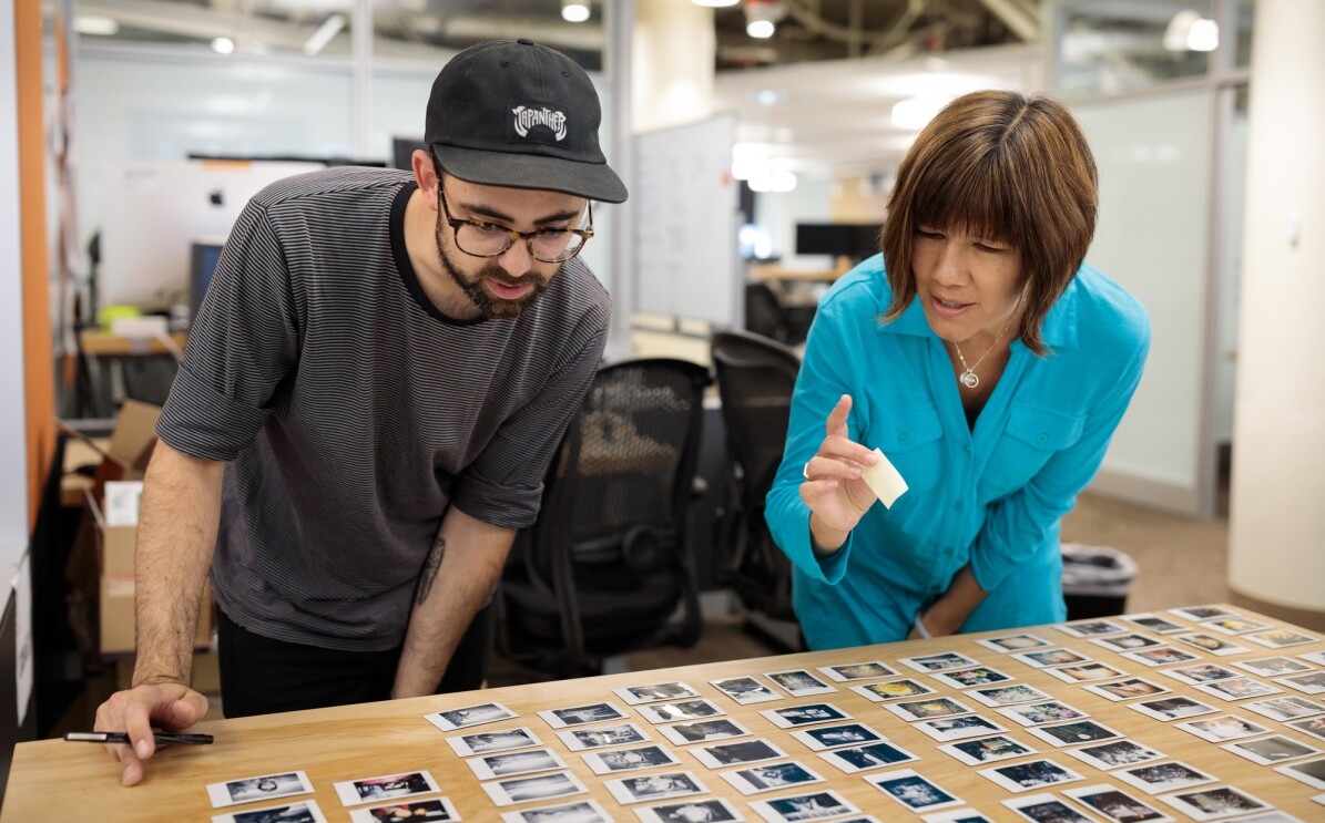 A man in a baseball cap and a woman in a teal blue top lean over a desk in an office setting. On the desk are dozens of small photographic prints. The woman holds a sticky note in her hand and prepares to point at an image.