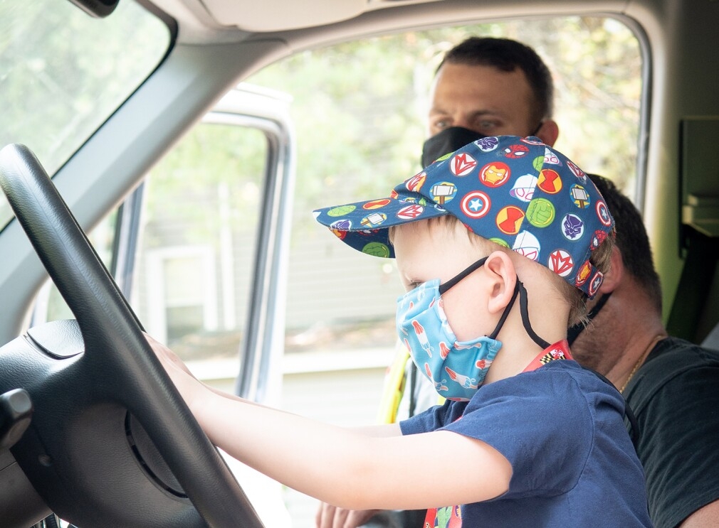 An image of a boy in the driver's seat of a van. He is pretending to drive and sitting next to his father. Both are wearing masks. 