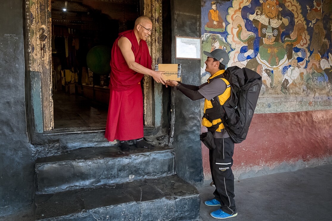 A Buddhist monk in glasses and maroon-colored robes accepts an Amazon package from a uniformed delivery associate. The associate stands at street level and is flanked by a colorful mural. The monk stands in a doorway on the second of three steps that lead into the building behind him.
