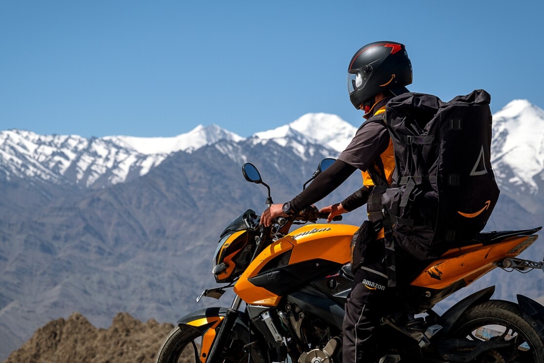 A helmeted motorcyclist wears a large black backpack with the Amazon smile logo. Snow-capped mountain peaks are in the background of the image.
