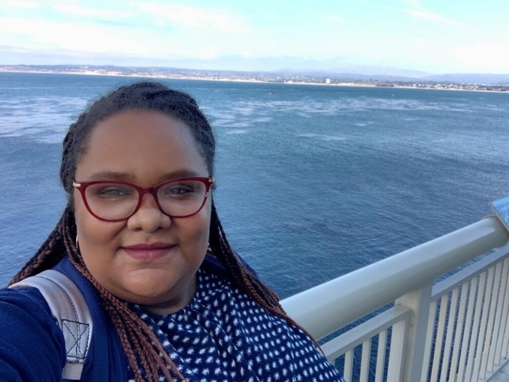 A woman smiles at the camera, with a smile on her face, in front of the ocean. 