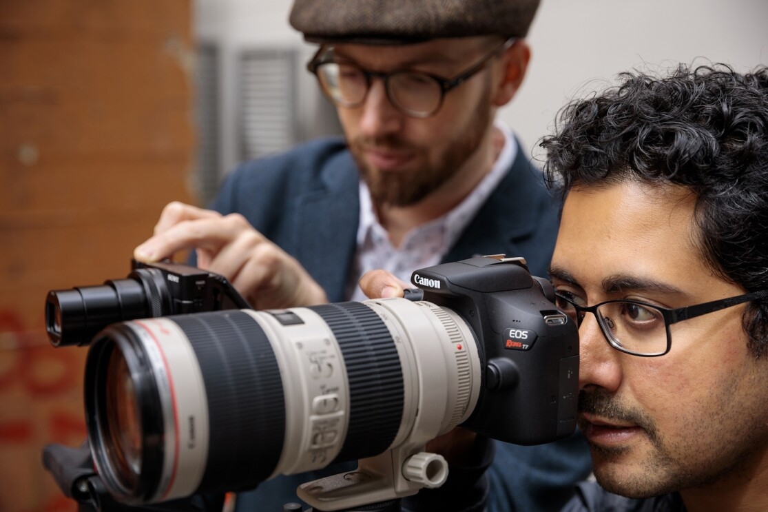 A man wearing eyeglasses uses a digital single-lens reflex camera equipped with a large professional lens. A second man is out of focus in the background.