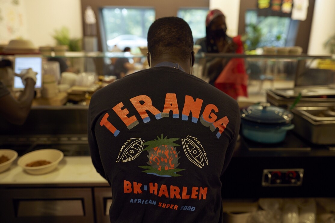 An image of a man working at the counter of a restaurant. You can see the back of his shirt and it reads "TERANGA BK HARLEM AFRICAN SUPER FOOD."