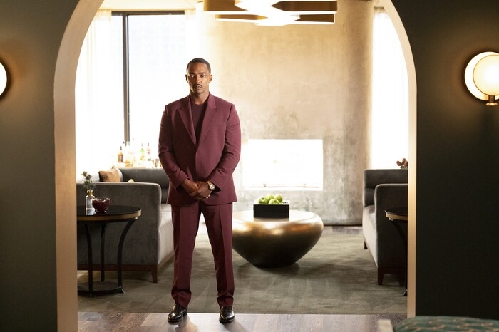 An image of Anthony Mackie standing in a doorway with his hands folded in front of him and a serious expression on his face. He is wearing a maroon suit.
