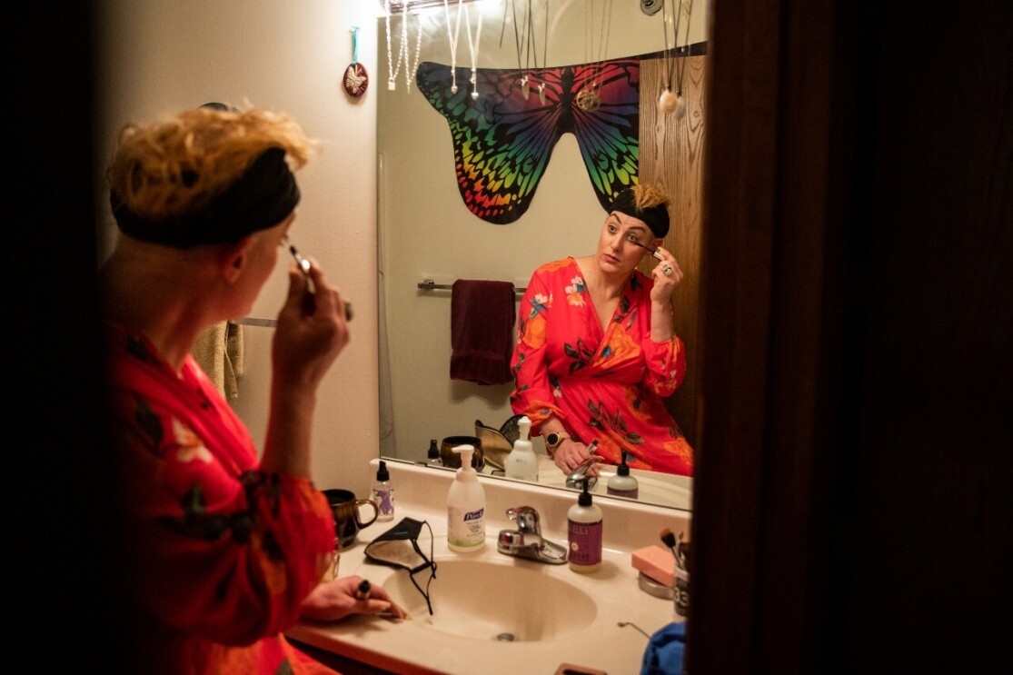 A woman sits in front of her bathroom mirror applying mascara.