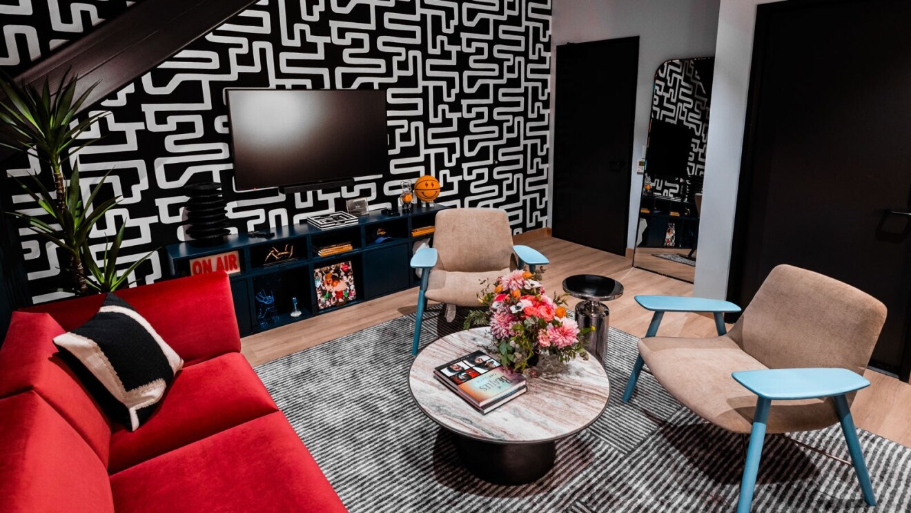 A room with a bright red couch, funky black and white wallpaper, a TV, a coffee table, and two blue chairs. It is one of the green rooms in the amazon music studio in los angeles.