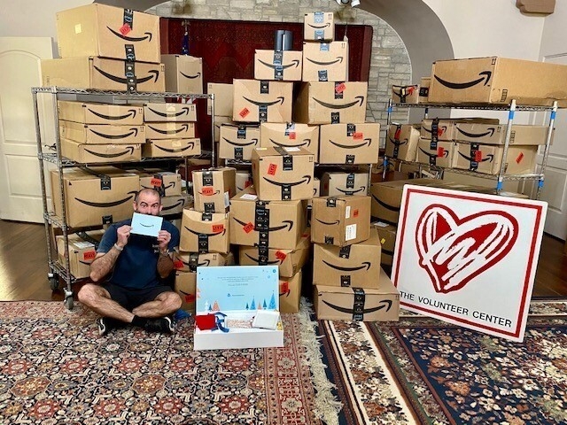 An image of Amazon boxes stacked up with a man sitting in front of the boxes holding the Delivering Smiles invitation for charities selected for this year's campaign. There's also a sign with a big heart and "The Volunteer Center" on it. 