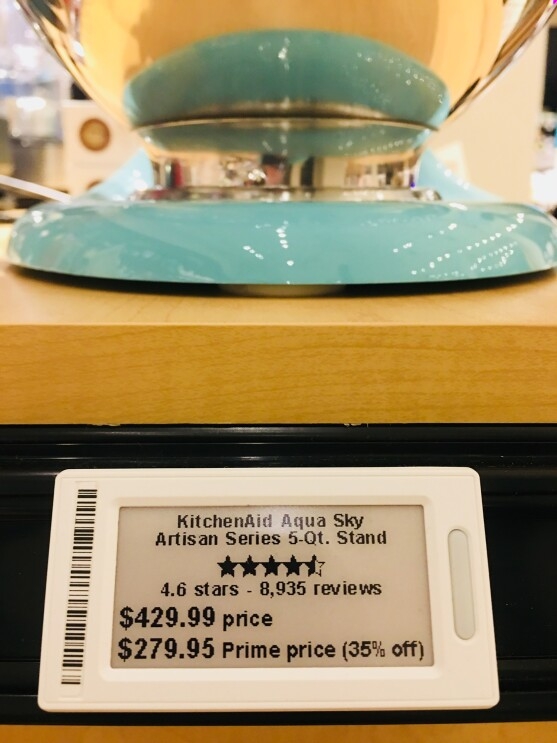 A digital tag for a KitchenAid Artisan Series 5-Qt. stand mixer in aqua sky. The tag shows that the mixer has 4.6 stars and 8,935 reviews at the time the photograph was taken. The regular price is $429.99, with a Prime member price of $279.95 (35 percent off). Behind the tag is an aqua blue KitchenAid stand mixer with stainless bowl.