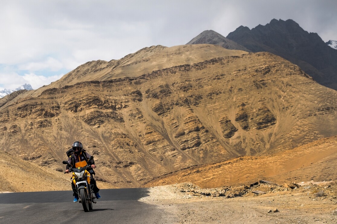 A motorcyclist rides on an asphalt road. Behind him, the road curves to the right and disappears from view. In the background a tan rock formation rises up from the road.