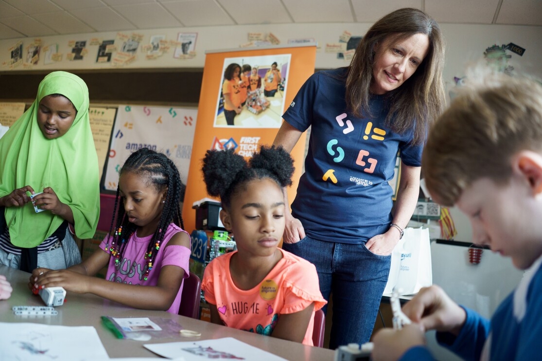 An Amazon employee wearing a Future Engineer t-shirt overlooks a group of students troubleshooting a device.