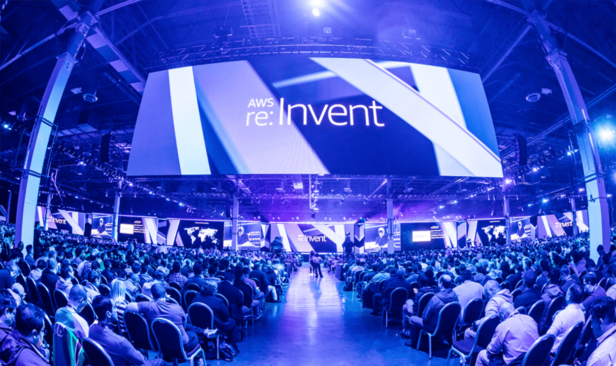 AWS re:Invent conference, view shows hundreds of individuals sitting in chairs at a conference. A huge screen hangs above them, the space is awash in blue light.