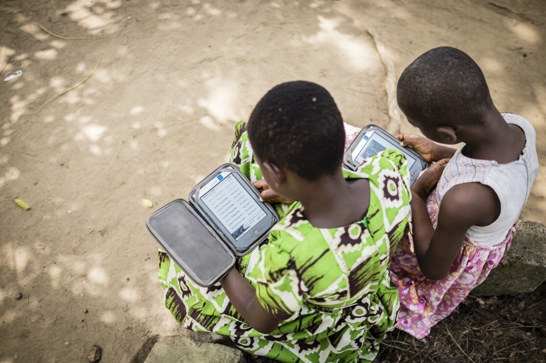 Two girls, one in a green dress and one in a pink and white dress, sit on a ledge and read from their Kindle Worldreaders.