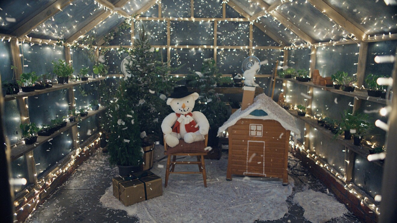 A lit up greenhouse with filled with a tree, stuffed animal, toy house, and fake snow.