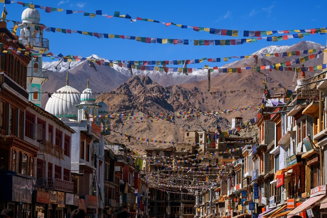 Under a bright blue sky and colorful prayer flags, multi-story buildings with shops are photographed on facing sides of a street. Domed structures and snow-capped mountains are in the background.