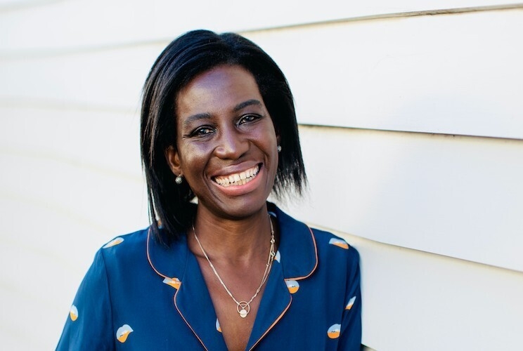 Prisca Ongonga-Daehn smiles for the camera standing outside. She wears a blue blouse with a gold necklace.