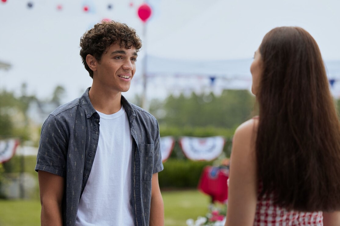 A young man stands in front of a young girl in an outdoor setting. He is smiling at her while she is talking. There are fourth of july decorations in the background. 