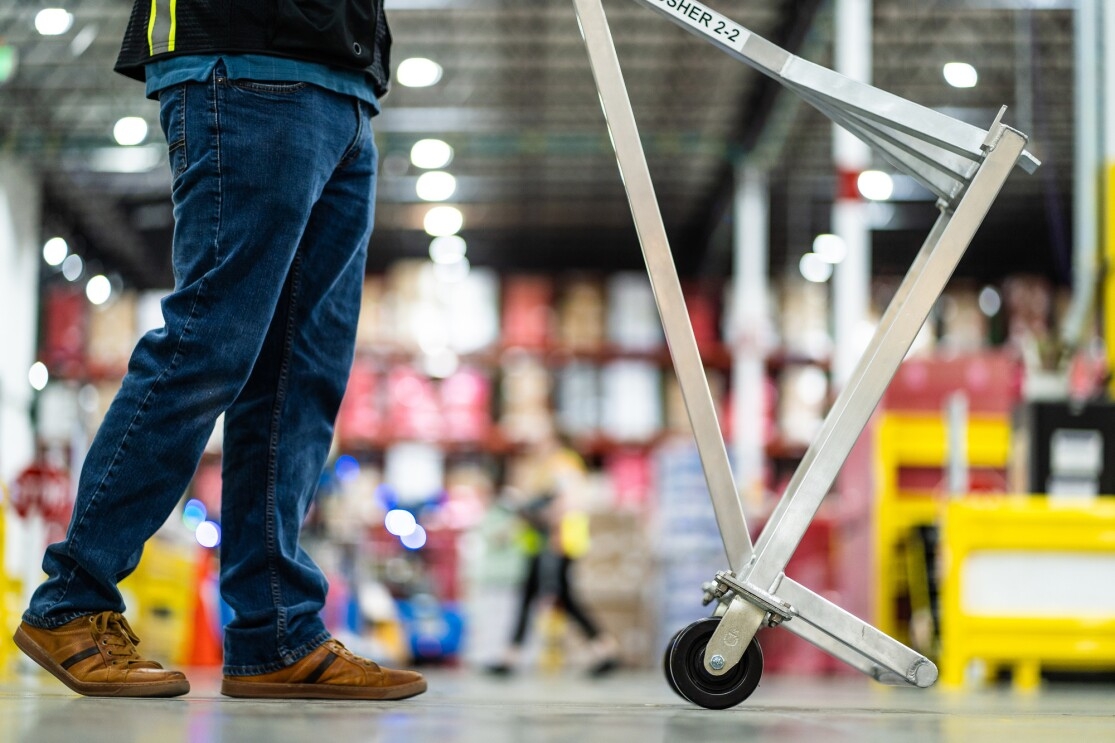 A man is photographed from the legs down. A metal frame on wheels in directly in front of him.