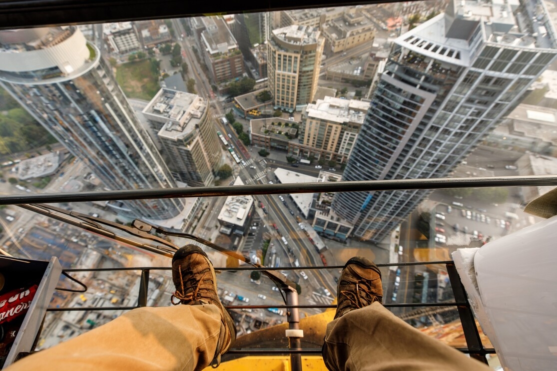 A man in a khaki pants and lace up shoes stands atop a crane, high above Seattle, WA.
