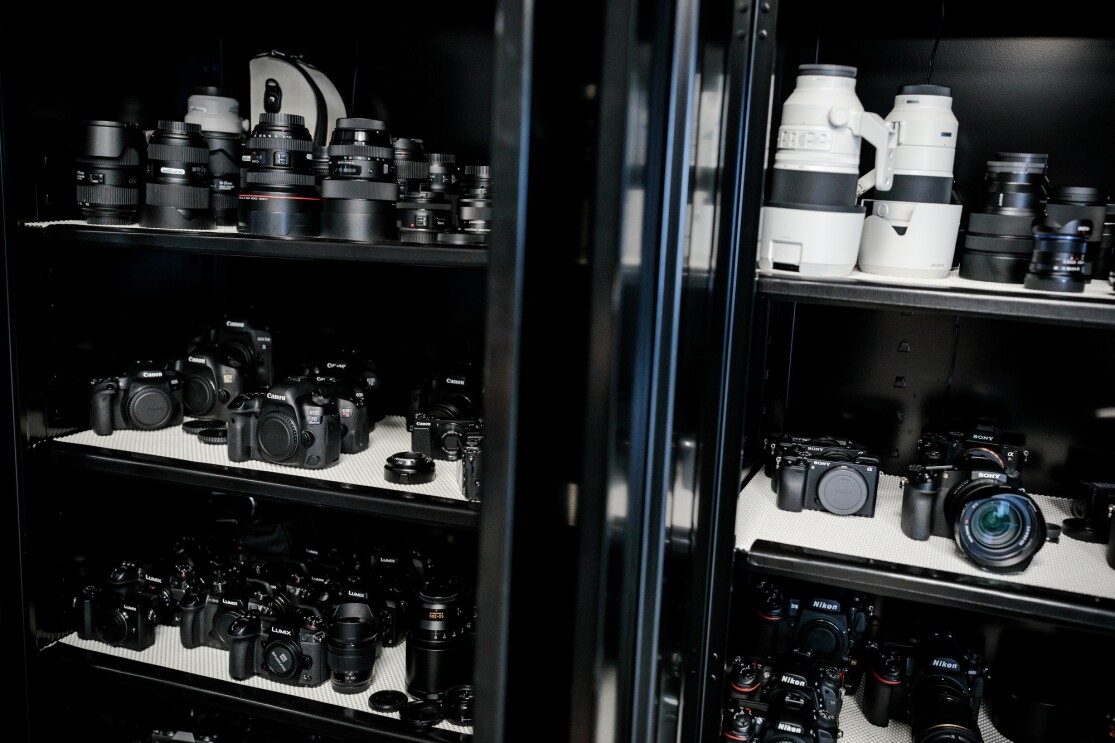 A closed metal cabinet with glass doors holds dozens of cameras and lenses.