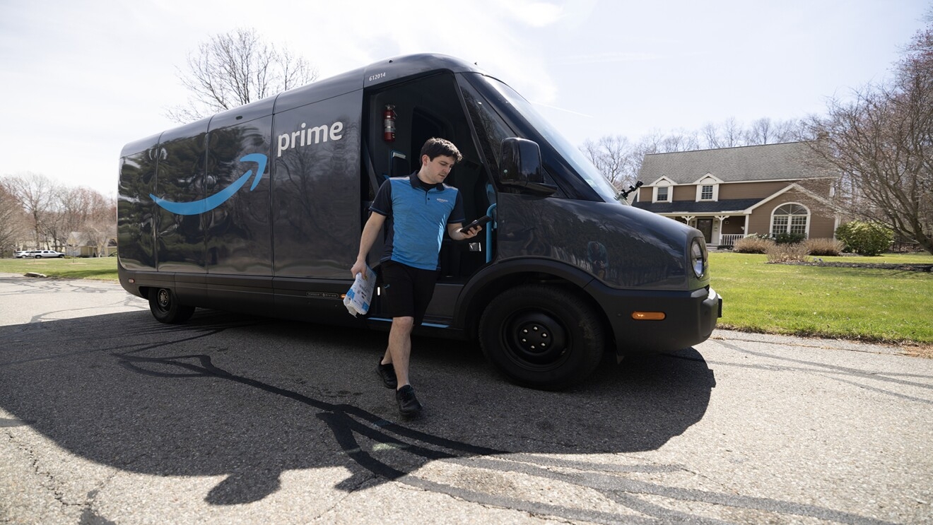 An Amazon delivery driver walks away from a Rivian van holding a package to deliver to a customer's home.