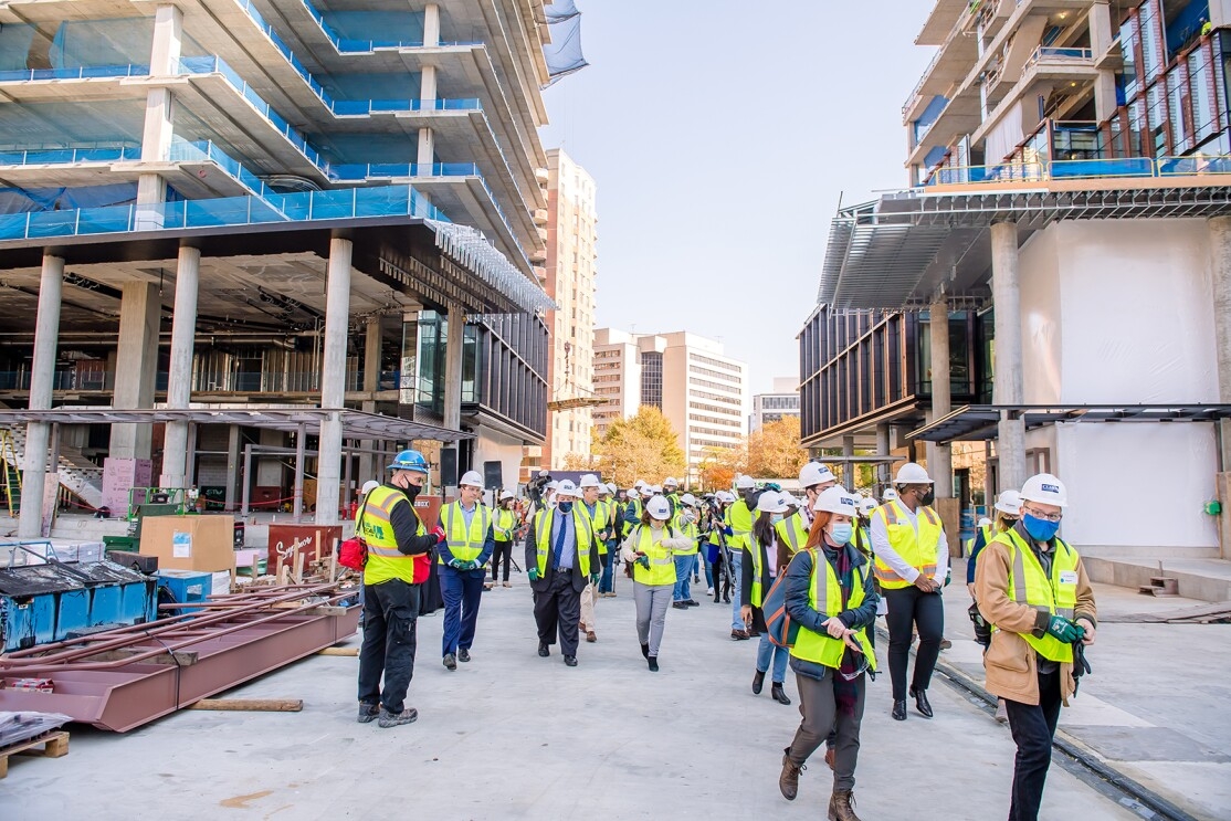 An image of people touring the construction site of Amazon's second headquarters while wearing hard hats that say "Amazon" and bright yellow safety vests.