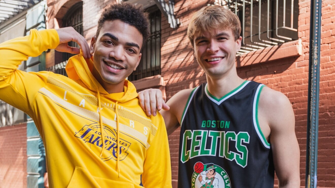 An image of two men standing in front of a red brick building. One is wearing a yellow hoodie that says "Lakers" on it in white writing with the Lakers logo below it. The other is wearing a black Boston Celtics jersey with the Celtics logo on it and lettering that says "Celtics."