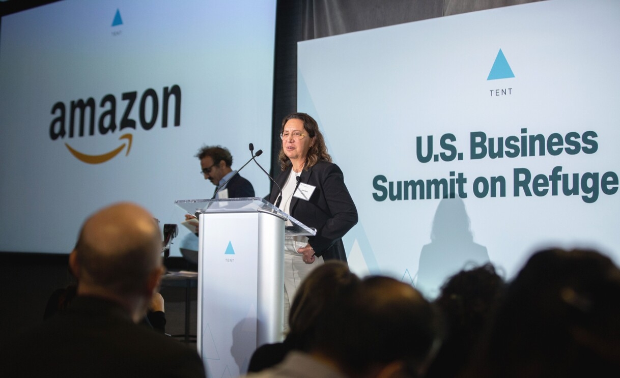 Janet Saura, VP of Employee Relations, standing at a podium announcing Amazon's hiring commitment at the U.S. Business Summit on Refugees.