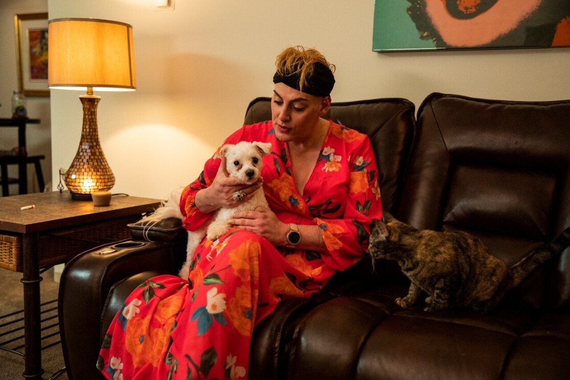 An image of a woman in a pink, floral robe relaxing on her couch with a small white dog and a brown and gold cat. 