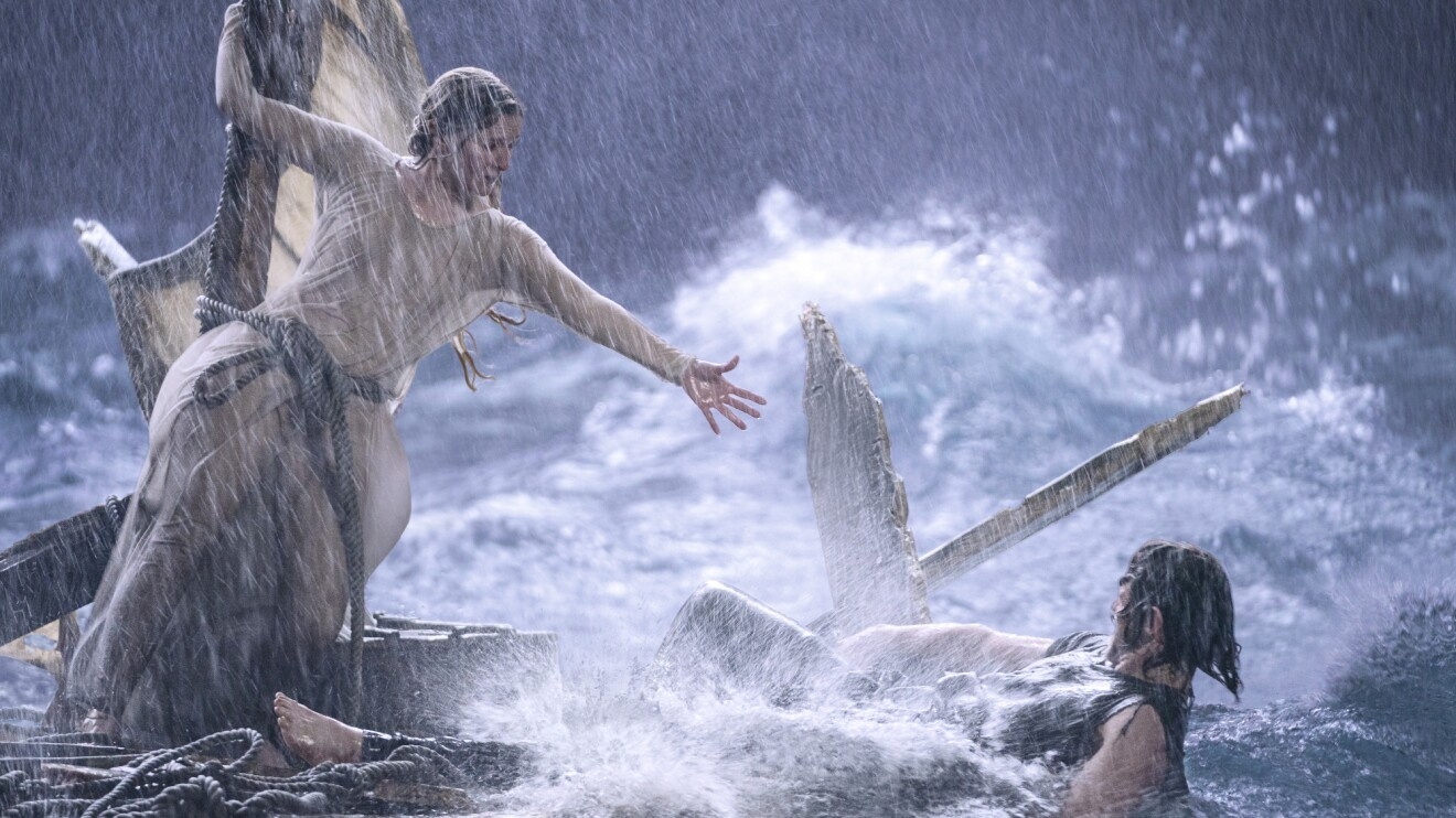 A man and a woman are pictured braving a storm in the ocean, causing their boat to break and the man falling in the water while the woman reaches for him.