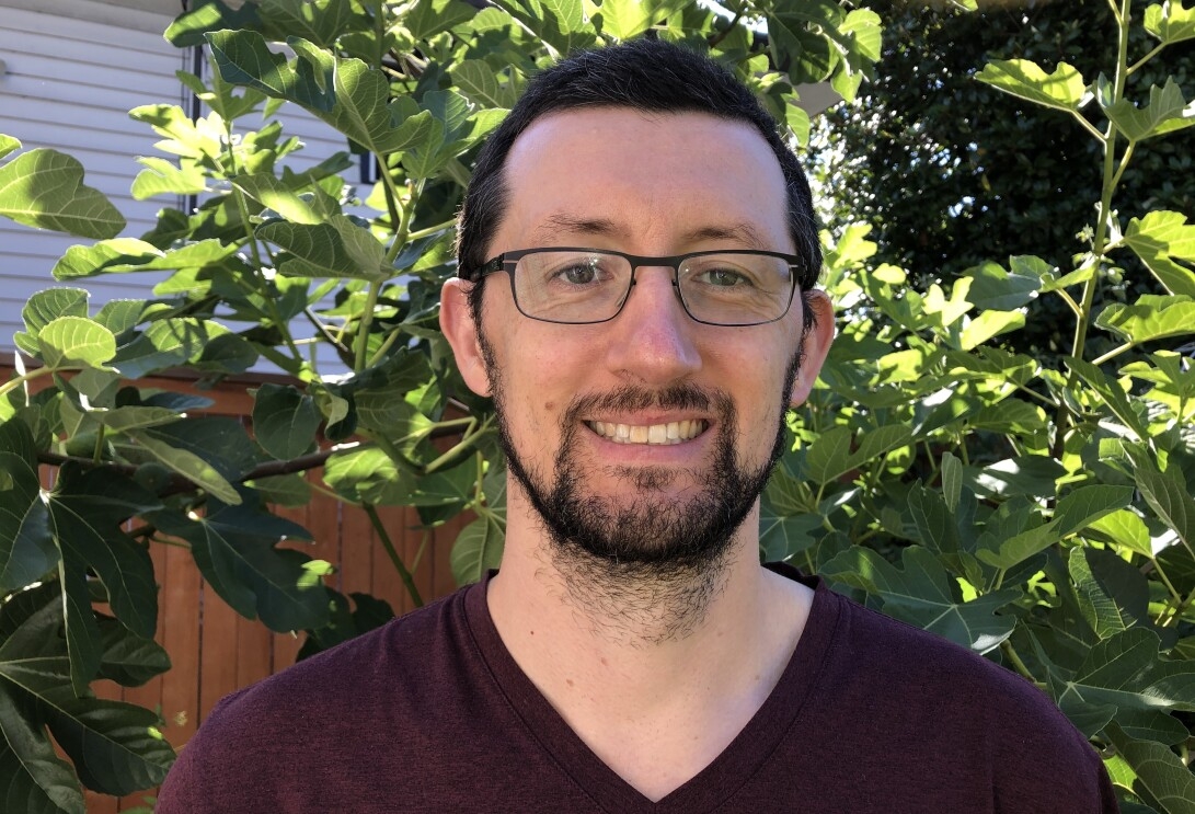 A man with glasses smiles at the camera. Behind him, greenery grows in an outdoor setting. 