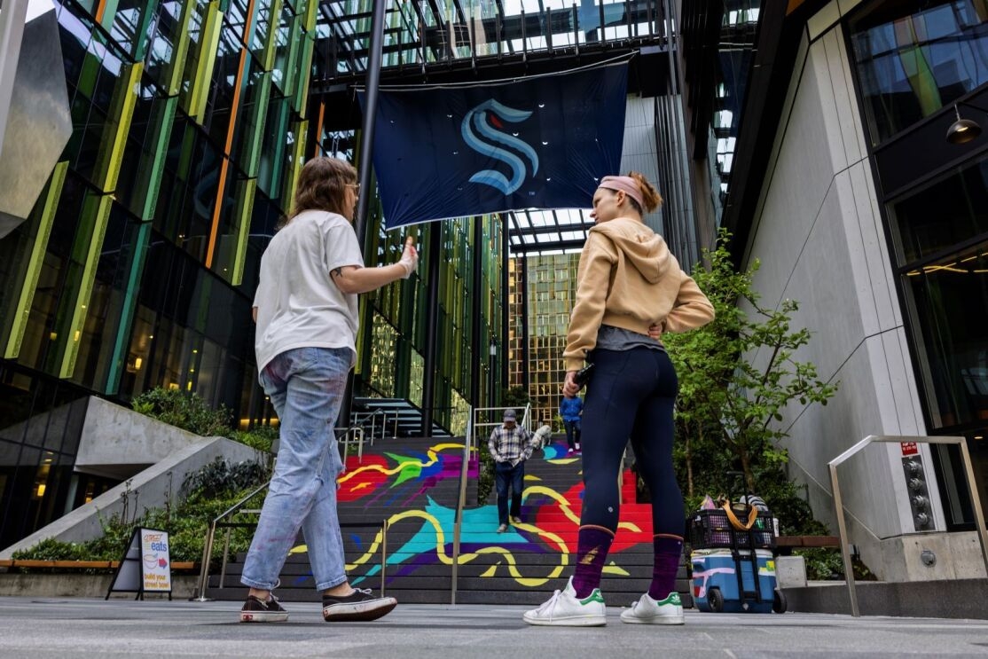 Two women in casual clothes hold a conversation. Behind them a large Seattle Kraken flag is hanging between two downtown buildings