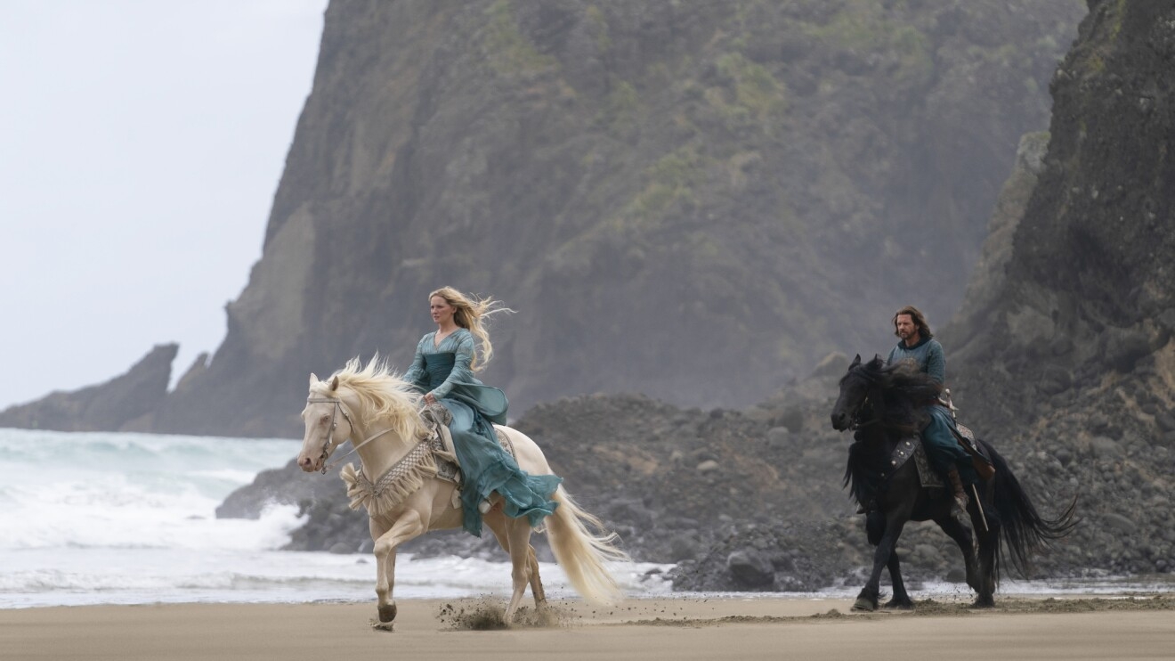 Two characters ride horses along a beach with cliffs in the background; the woman rides a white horse while the man rides a black horse.