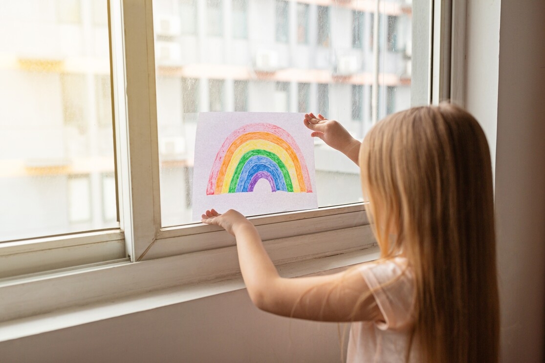 A child placing homemade rainbows in the window of their home, during COVID-19 pandemic.