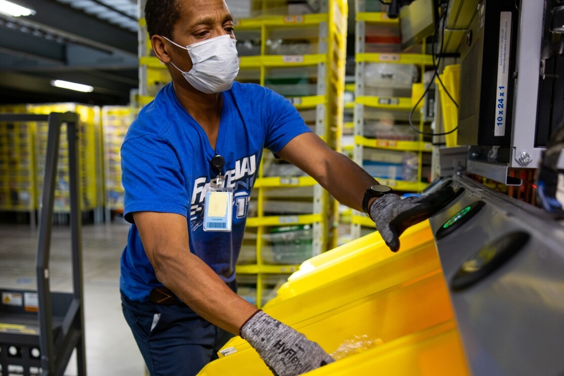 A man works while wearing gloves and a mask that covers his nose and mouth. He reaches for an illuminated button and handles a yellow bin.