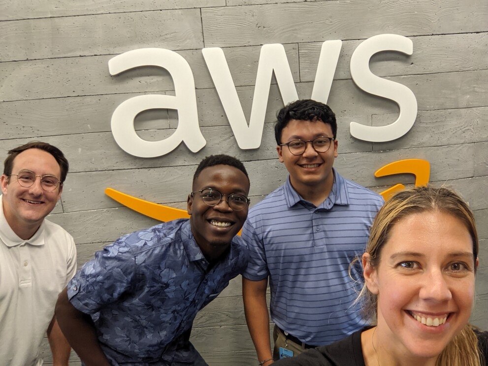A woman holds the phone for a group selfie in front of the AWS logo on a wall in an Amazon office.