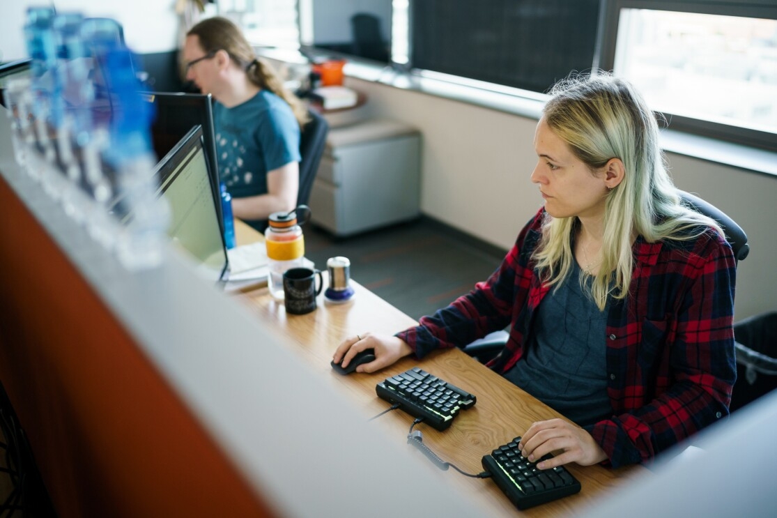 Danielle Skysdottir sits at her desk in a cubicle at Amazon's Seattle headquarters. She has blond hair and is wearing a T-shirt and flannel shirt. She's using a computer mouse with her right hand and typing on a split ergonomic keyboard with her left hand.