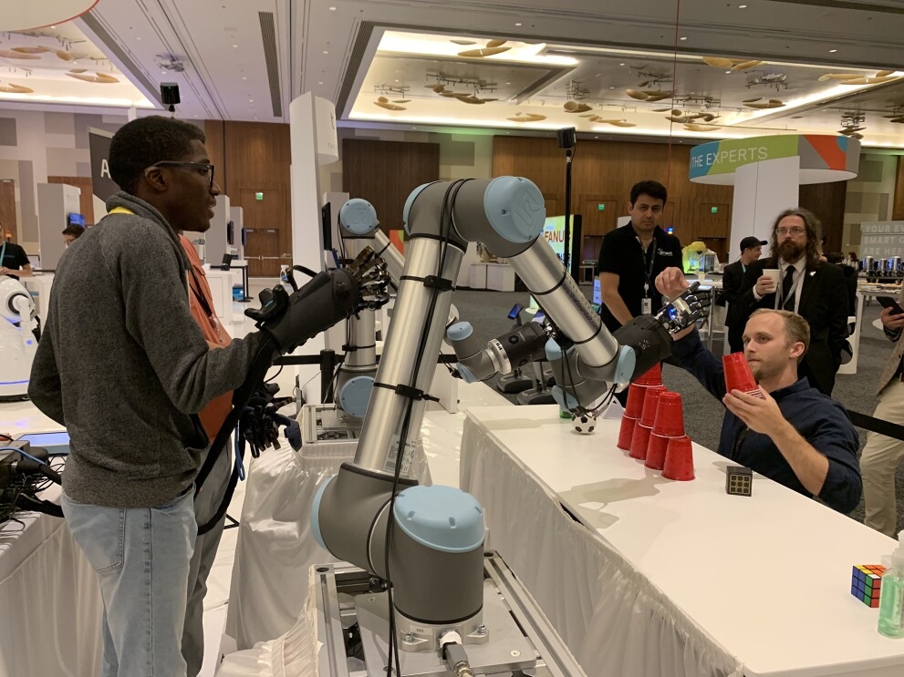 A teen, Leo Jean Baptiste, wears robotic-control gloves and controls a pair of robotic arms with his motions.