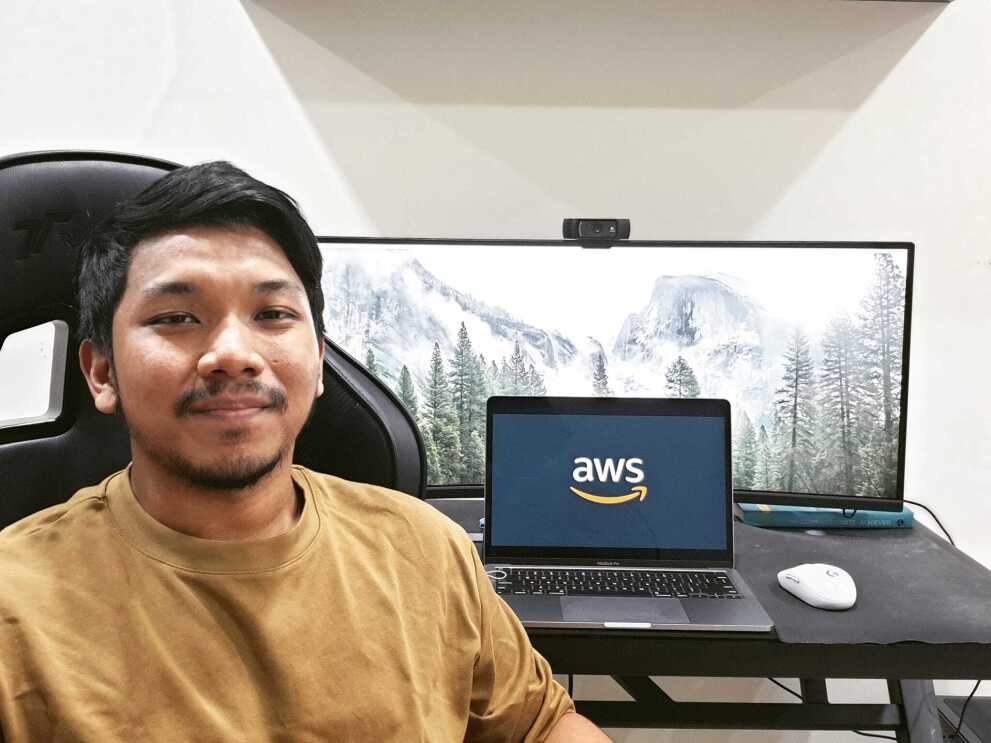 An image of a man smiling for a photo while sitting at his desk in his home. His laptop monitor in the background has the Amazon logo on it. 
