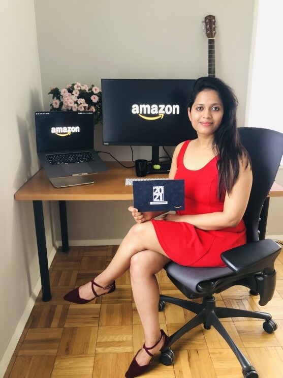 An image of a woman smiling for a photo while sitting at her desk in her home. She has two computer monitors in the background with the Amazon logo on their screens, and she is holding an invitation to the 2021 Amazon internship class.