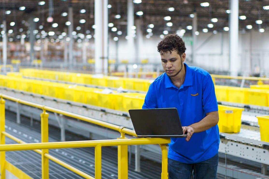 A man in a blue polo shirt works on a laptop in a large, high-ceilinged space. Yellow plastic totes are behind him and stretch into the distance.