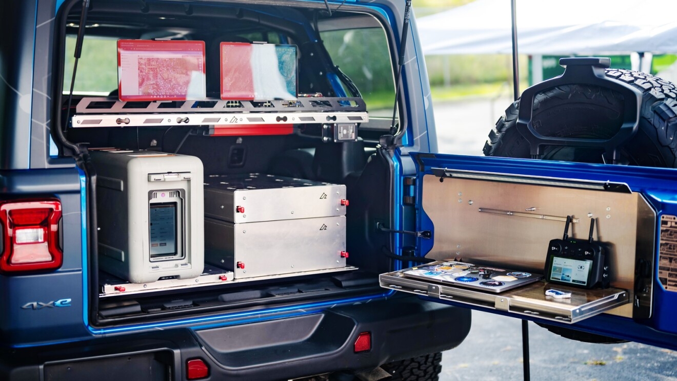 An image of the back of a blue Jeep with the door open. There are several pieces of computer equipment inside.