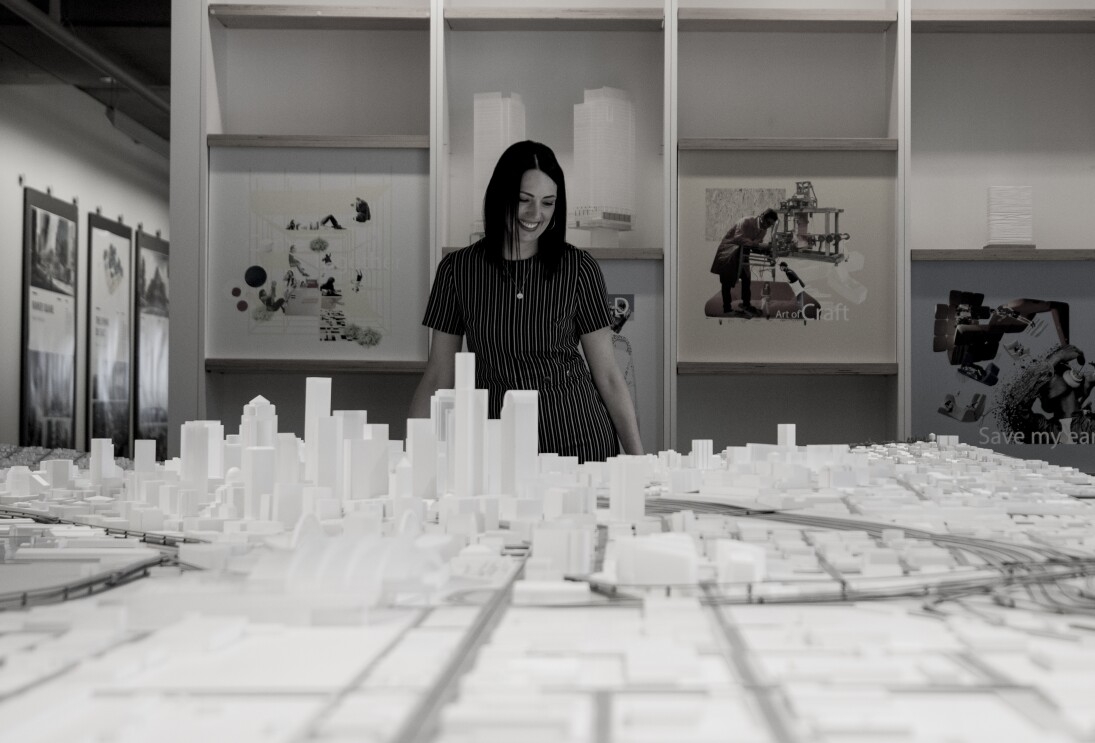 A woman in a grey-striped shirt stands over a large, scale model of downtown Seattle, looking down at the model.